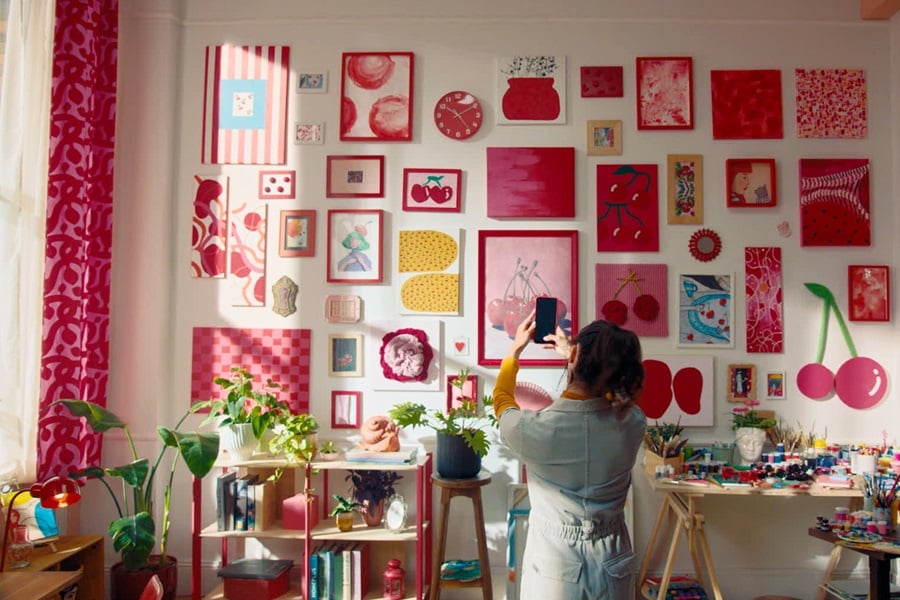 Sala de estar con arte de temática de cerezas colgado con tiras Command, además de un estante con libros y plantas y una mesa con materiales de arte.
