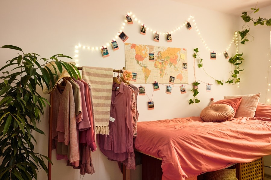 Pared de dormitorio estudiantil con luces decorativas, fotos, un mapa del mundo y ramas de plantas colgados con ganchos transparentes Command, además de una cama ordenada y un perchero.
