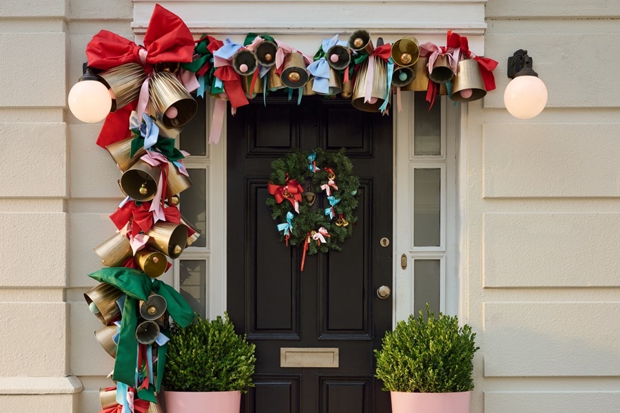 Puerta principal con una guirnalda de campanas doradas y cintas rojo‑verdes colgada con ganchos Command Outdoor, además de una corona en un gancho Command para coronas.
